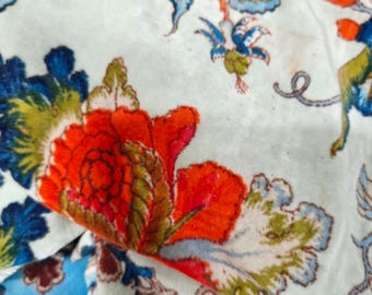 May include: Close-up of a floral fabric with a cream background. The design features large, vibrant red flowers with green and orange accents, and blue and green leaves. The fabric has a soft, velvety texture.