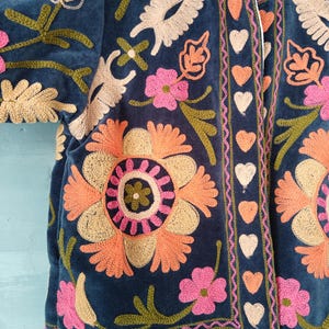 以下が含まれることがあります： ネイビーブルーの刺繍入りジャケット、花柄のデザイン。オレンジとピンクの大きな花、クリームとグリーンのアクセントが特徴です。縁はピンクと白のハートでトリミングされています。柔らかいベルベットのような素材で作られています。