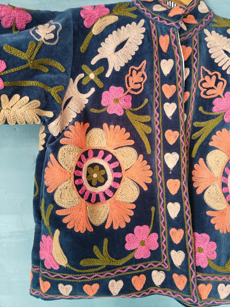 以下が含まれることがあります： ネイビーブルーの刺繍入りジャケット、花柄のデザイン。オレンジとピンクの大きな花、クリームとグリーンのアクセントが特徴です。縁はピンクと白のハートでトリミングされています。柔らかいベルベットのような素材で作られています。