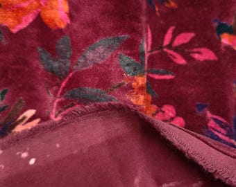 May include: Close-up of a burgundy velvet fabric with a floral pattern. The flowers are in shades of pink, orange, and purple, with green leaves. The fabric has a soft, textured appearance, with a contrasting burgundy underside.