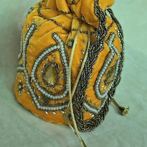 Könnte beinhalten: Senfgelber Samtbeutel mit Kordelzug und aufwendiger Perlenarbeit. Die Tasche hat weiße und silberne Perlen, die tropfenförmige Formen umreißen, und einen braunen Perlenrand. Ein goldener Kordelzug und dekorative Quasten vervollständigen das Design.