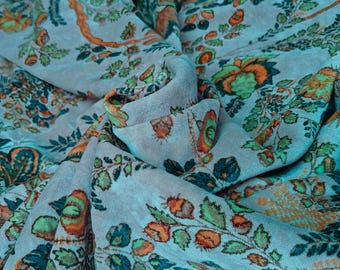 May include: Close-up of a light blue fabric with a floral pattern in shades of orange, green, and teal. The fabric is draped, showing the texture and design of the material. The design includes leaves, flowers, and other botanical elements.
