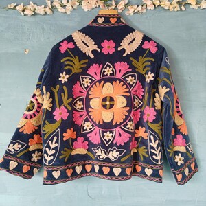 以下が含まれることがあります： ネイビーブルーのベルベットジャケット、カラフルな花の刺繍入り。 ジャケットの背面には、ピンク、オレンジ、クリーム色の花を使った大きな円形のフラワーデザインが施されています。 袖と襟にも、花の刺繍とハートの模様が施されています。