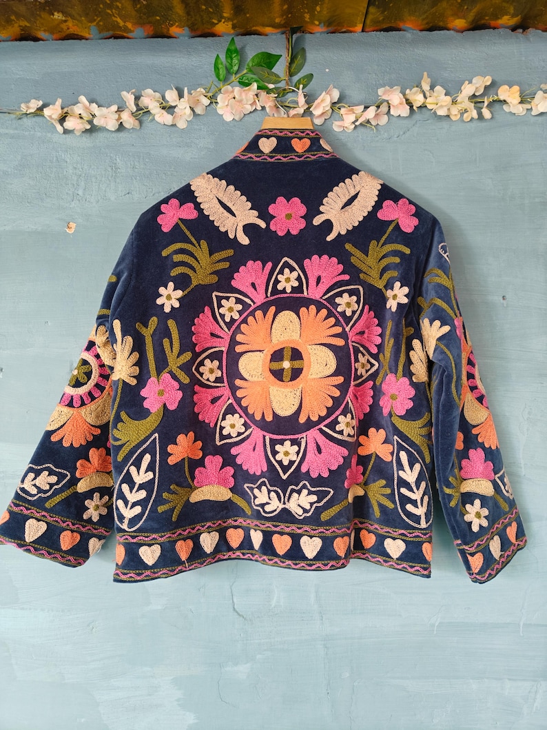 以下が含まれることがあります： ネイビーブルーのベルベットジャケット、カラフルな花の刺繍入り。 ジャケットの背面には、ピンク、オレンジ、クリーム色の花を使った大きな円形のフラワーデザインが施されています。 袖と襟にも、花の刺繍とハートの模様が施されています。