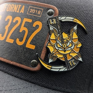 May include: A black baseball cap with a yellow and black license plate that says "FORNIA 2018 3252" and a yellow and black enamel pin with a rose, scythe, and the letters "LA FC".
