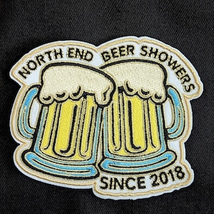 May include: White embroidered patch with two yellow beer mugs with blue and gold trim. The text "North End Beer Showers Since 2018" is embroidered in gold on the patch.