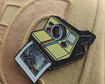 Puede incluir: Un pin de esmalte amarillo y negro de una cámara vintage con una foto insertada. La foto muestra una imagen borrosa de un cielo nocturno con estrellas.