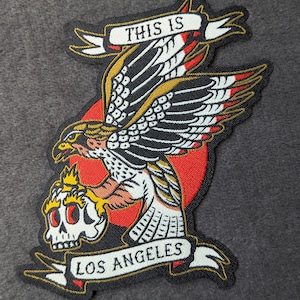 May include: A black and white embroidered patch with a red, white, and gold eagle holding a skull in its talons. The eagle is flying over a red sun. The patch has a banner that says "This is Los Angeles".
