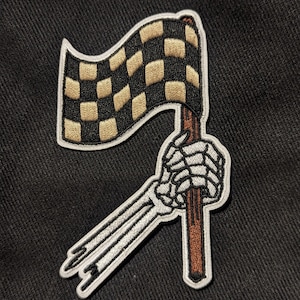 May include: A black and white checkered flag held by a skeleton hand with a brown stick. The flag is waving in the wind.