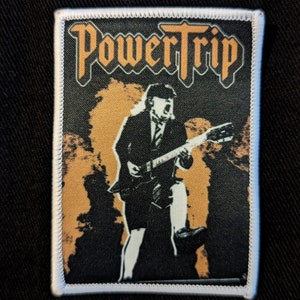 Puede incluir: Un parche bordado en blanco y negro con el texto "Power Trip" en una fuente estilizada. El parche presenta la silueta de una persona tocando una guitarra frente a un fondo de llamas amarillas y naranjas.