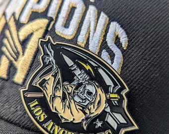 Puede incluir: Una gorra de béisbol negra con bordado dorado y blanco que dice "MLS Cup Champions". Un pin de esmalte dorado y negro con un diseño de calavera y guadaña está sujeto a la parte delantera de la gorra. El pin dice "Los Angeles FC".