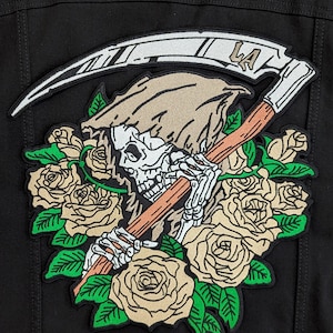 Puede incluir: Un parche bordado en blanco y negro que representa un esqueleto sosteniendo una guadaña con la palabra "LA" en ella. El esqueleto lleva una túnica marrón con capucha y está rodeado de rosas blancas con hojas verdes.