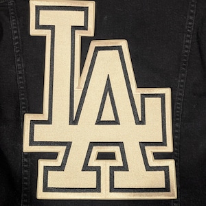 Peut inclure: Veste en jean noire avec un grand écusson brodé "LA" beige et noir dans le dos. L'écusson présente un design de lettres en blocs audacieux, mettant en valeur le logo emblématique.