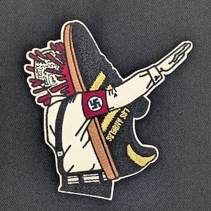 May include: An embroidered patch featuring a cartoon figure in a white shirt and black pants, giving a Nazi salute. The figure is holding a black and yellow shoe with the text "STADIUM 97" on the side. The figure is wearing a red armband with a white swastika.