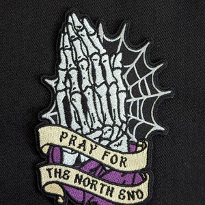 May include: A black and white embroidered patch with a skeleton hand in a praying position. The hand is surrounded by a spider web and a banner that reads "Pray for the North End".