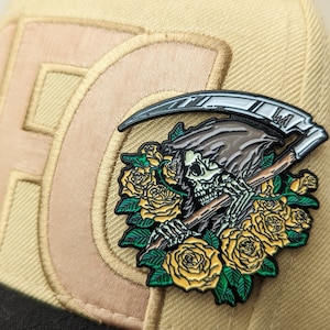 May include: A black and silver enamel pin featuring a skeleton figure holding a scythe, surrounded by yellow roses with green leaves. The pin is attached to a beige baseball cap.