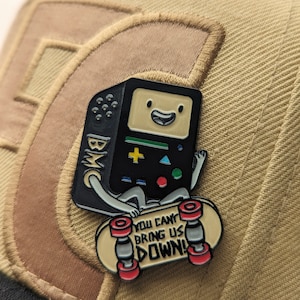 May include: A black and green enamel pin of a cartoon character, resembling a retro video game console, riding a skateboard. The character has a smiling face and is wearing a black hat. The skateboard is yellow with red wheels and the text "You can't bring us down!" printed on it.