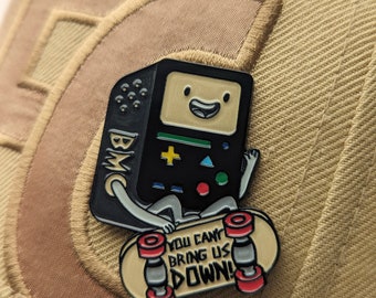 Puede incluir: Un pin de esmalte negro y verde de un personaje de dibujos animados, que se asemeja a una consola de videojuegos retro, montando un monopatín. El personaje tiene una cara sonriente y lleva un sombrero negro. El monopatín es amarillo con ruedas rojas y el texto "You can't bring us down!" impreso en él.