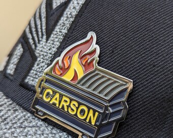 Puede incluir: Una gorra de béisbol negra y gris con un pin de metal que presenta una llama amarilla, naranja y roja en la parte superior de un contenedor de basura azul con la palabra "CARSON" en letras amarillas.