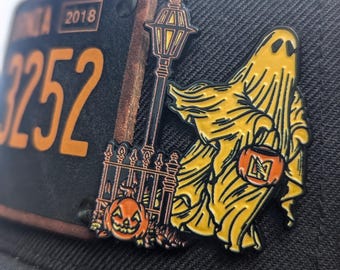 Puede incluir: Una gorra de béisbol negra con un pin de metal que representa un fantasma sosteniendo una bolsa de truco o trato, una farola, una valla y una calabaza tallada. El pin tiene una placa de matrícula vintage con el número 3252 y el año 2018.