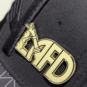 May include: A gold-colored metal pin with the letters "AFD" in a stylized font. The "A" is formed by a hand with two fingers pointing up. The pin is attached to a black fabric surface.
