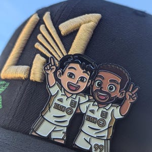 May include: A black baseball cap with gold embroidered letters and a pin of two cartoon soccer players in white and gold uniforms. The pin features the text "BMO" and the number "99".