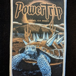 May include: A black and white embroidered patch with the text "Power Trip" in a bold font. The patch features a desert scene with a spiky turtle and the text "Indio, CA 2023".