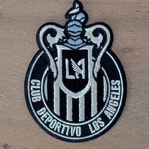 Puede incluir: Parche bordado con el logo del Club Deportivo Los Ángeles. El parche tiene una combinación de colores negro y plateado con el escudo del equipo en el centro. El texto "CLUB DEPORTIVO LOS ANGELES" está bordado alrededor del borde.