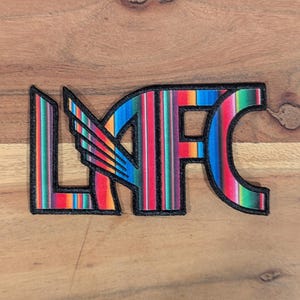 May include: A colorful LAFC patch with a black border. The letters LAFC are filled with a striped pattern of red, orange, green, blue, and yellow. A wing design is on the left side of the letters.