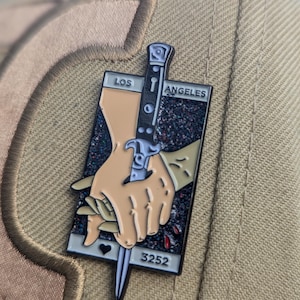 May include: A black and white enamel pin with a black knife and two hands holding it. The pin has a black background with glitter and red blood drops. The text "LOS ANGELES" and "3252" are on the pin.