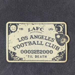 May include: A beige and black embroidered patch with the text "LAFC", "Los Angeles Football Club", "0003252000", and "Til Death". The patch features a crescent moon, a sun, and an eye.