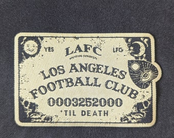 Puede incluir: Un parche bordado beige y negro con el texto "LAFC", "Los Angeles Football Club", "0003252000" y "Til Death". El parche presenta una luna creciente, un sol y un ojo.