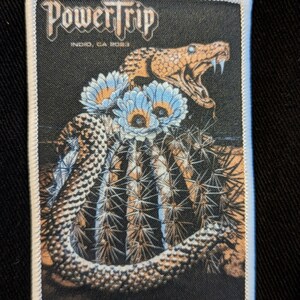 Puede incluir: Un parche bordado en blanco y negro con el texto "Power Trip" y las palabras "Norco, CA 2023". El parche presenta una imagen detallada de una serpiente envuelta alrededor de un cactus con flores azules.