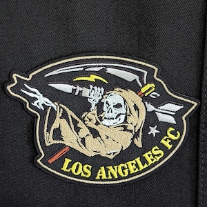 May include: A black and white embroidered patch with a skeleton wearing a hooded robe holding a scythe. The patch has a yellow star and the text "Los Angeles FC" in yellow.