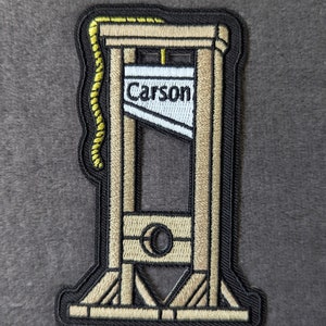 May include: A black and beige embroidered patch of a guillotine with the word "Carson" on the blade.