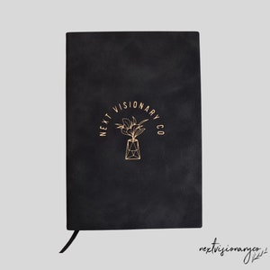 Op de afbeelding: Een zwart notitieboek met een gouden logo dat "Next Visionary Co." zegt en een gestileerde afbeelding van een plant in een pot.