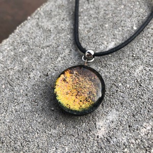 Könnte beinhalten: Eine schwarze Schnurkette mit einem runden, irisierenden Glasanhänger. Der Anhänger hat eine dunkelgrüne Basis mit goldenen und orangen Flecken.