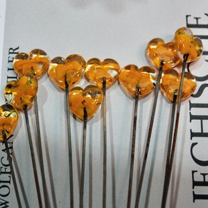 May include: Close-up of several heart-shaped glass ornaments on metal stems. The glass hearts are a translucent amber color. The stems are thin and metallic. The ornaments are arranged on a white surface with text in the background.