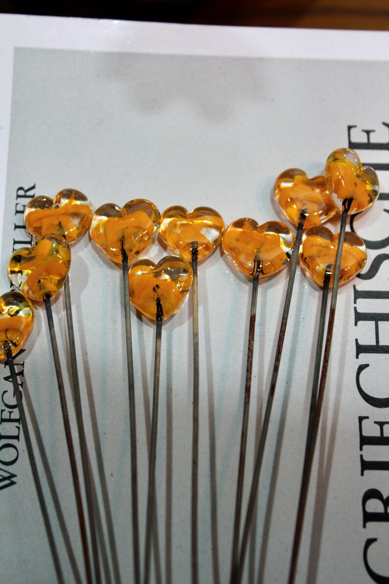 May include: Close-up of several heart-shaped glass ornaments on metal stems. The glass hearts are a translucent amber color. The stems are thin and metallic. The ornaments are arranged on a white surface with text in the background.