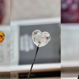 May include: Three heart-shaped lollipops on black sticks. One is amber with an orange design inside, one is clear with a white design, and the third is red. The lollipops are in front of a blurred background.