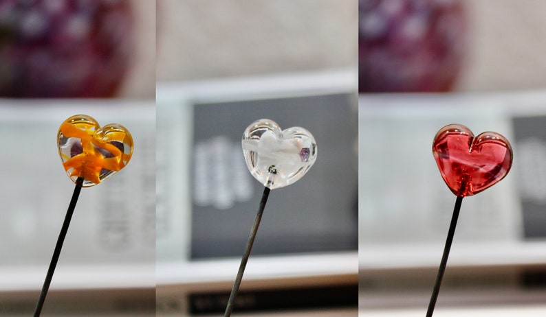 May include: Three heart-shaped lollipops on black sticks. One is amber with an orange design inside, one is clear with a white design, and the third is red. The lollipops are in front of a blurred background.