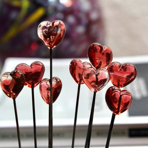 May include: A collection of glass heart-shaped ornaments in varying sizes. Each heart is a translucent red color and is attached to a thin, dark stem. The hearts are arranged in a cluster, creating a romantic and decorative display.