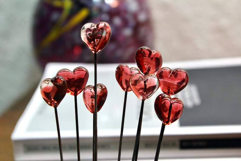 May include: A collection of glass heart-shaped ornaments in varying sizes. Each heart is a translucent red color and is attached to a thin, dark stem. The hearts are arranged in a cluster, creating a romantic and decorative display.