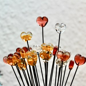 May include: A collection of heart-shaped glass ornaments in various colors, including clear, amber, and red. Each heart is mounted on a thin, black stem, creating a bouquet-like arrangement. The background is a blurred, light-colored surface.