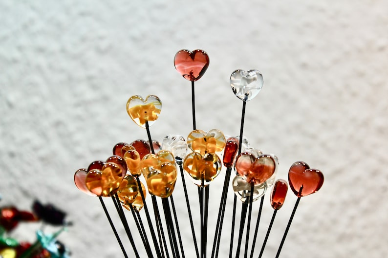 May include: A collection of heart-shaped glass ornaments in various colors, including clear, amber, and red. Each heart is mounted on a thin, black stem, creating a bouquet-like arrangement. The background is a blurred, light-colored surface.