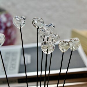 May include: A collection of clear, heart-shaped glass ornaments on thin black stems. The hearts are clustered together, creating a delicate and romantic display. The background is blurred, suggesting a soft focus on the decorative elements.