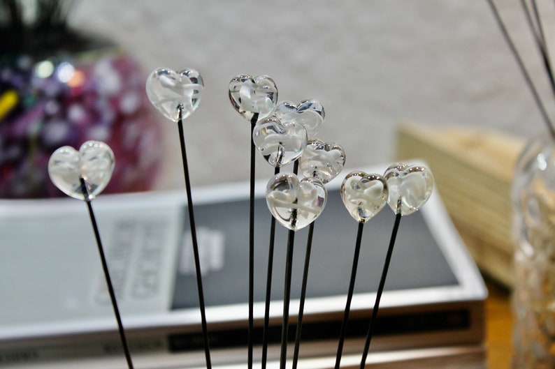 May include: A collection of clear, heart-shaped glass ornaments on thin black stems. The hearts are clustered together, creating a delicate and romantic display. The background is blurred, suggesting a soft focus on the decorative elements.