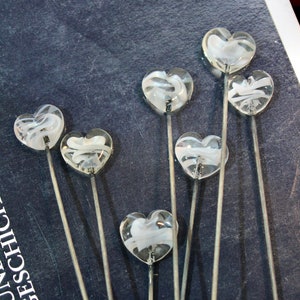 May include: A collection of clear glass heart-shaped decorative picks with white swirling designs. Each heart is mounted on a thin, silver-colored metal stem. The picks are arranged against a dark blue background.
