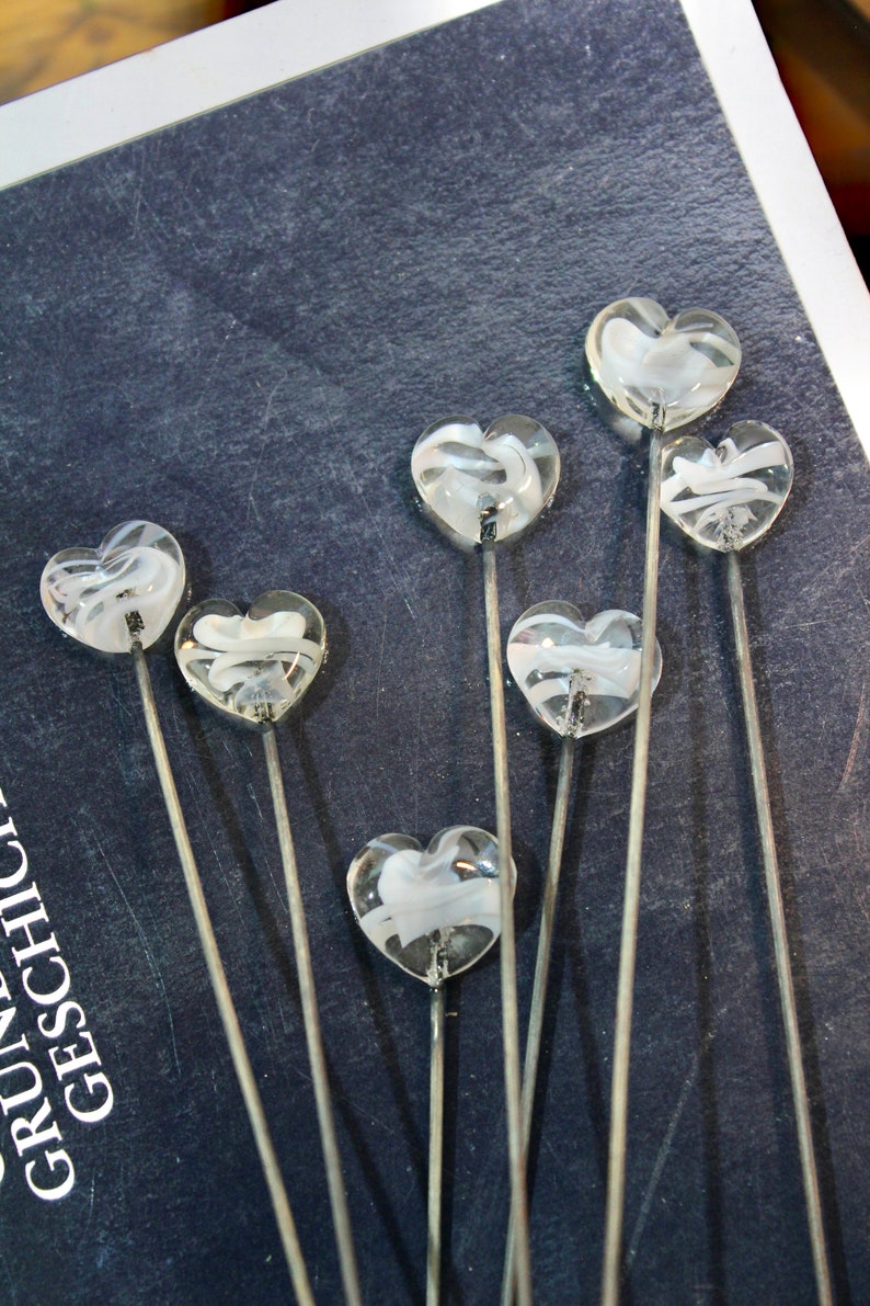 May include: A collection of clear glass heart-shaped decorative picks with white swirling designs. Each heart is mounted on a thin, silver-colored metal stem. The picks are arranged against a dark blue background.