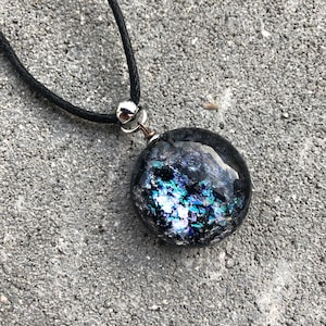 Könnte beinhalten: Eine schwarze Schnurkette mit einem silbernen Verschluss und einem runden, irisierenden Glasanhänger. Der Anhänger hat eine dunkelblaue Basis mit silbernen und blauen Sprenkeln.